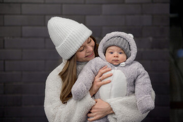 young woman with a baby in her arms walking around the city. mom and son are walking in cold weather, dressed in trendy warm clothes. Mom gently hugs her child.