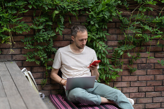 Concentrated serious man entrepreneur with laptop and phone sits outdoors near brick wall. Carried away guy runs successful business via Internet, wanting to find investments for own startup company