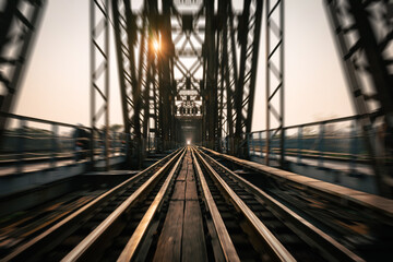 Train tracks lead downwards over a long metal bridge with a radial blur. There is a warm evening atmosphere.