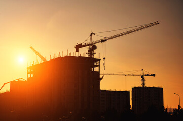 silhouette of construction crane building a house on a construction site against background of sunset orange sky