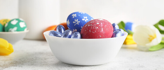 Bowl with painted Easter eggs and candies on light background