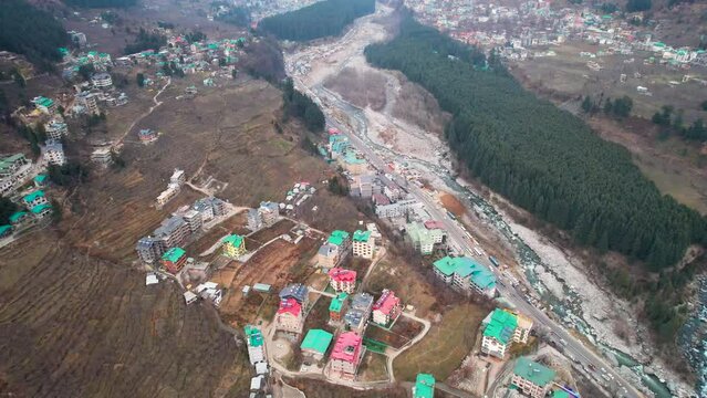 aerial drone shot of kullu manali showing the barren, rural areas of hill station with colorful houses, busy highway on side of beas river with forest and himalaya mountains