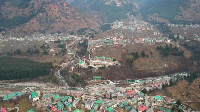 aerial drone shot flying forward over colorful houses, busy kullu manali highway beas river and hamta village on himalaya mountain in distance