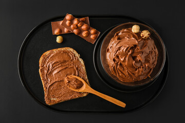Board with bowl of tasty hazelnut butter, toast and chocolate on dark background