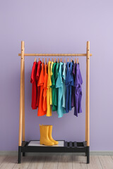 Rack with colorful t-shirts and gumboots near lilac wall