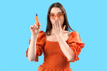 Shocked young woman in sunglasses with disposable electronic cigarette on blue background
