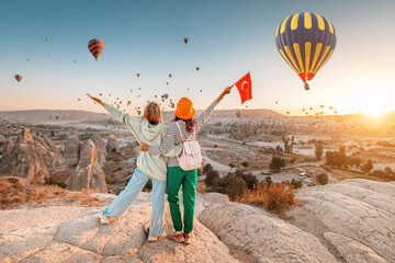 Girl friends travellers with turkish flag, hugging on a viewpoint and admiring view of flying hot...