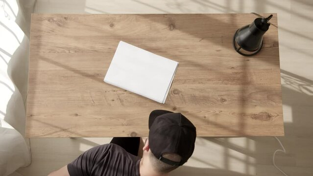 man in black cap sits down at table and write word START on white sheet of A4 paper with black marker. Top down view of guy at work desk, freelancer in coworking space. Beginnings.