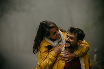 Happy affectionate couple in raincoats enjoying nature. A woman is jumping on her man and both having fun.