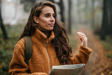 A pensive hiker woman walking in the forest, holding a map and wearing a backpack with camping gear.