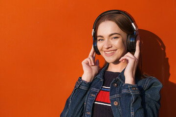 Pretty woman listening to music on headphones, outdoor hipster portrait against orange wall, wearing denim jacket