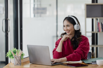 Happy smiling young asian business woman wear headphones working online sitting at desk in the office,