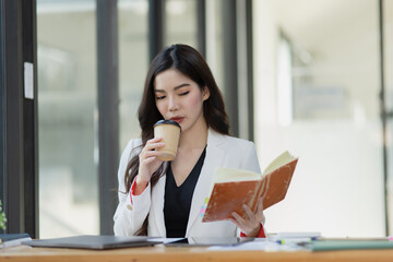 Friendly cheerful asian businesswoman reading a book and holding a coffee mug, people emotions and casual concept, positive woman working in office workspace.