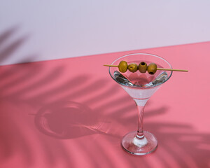 a martini glass with olives on a pink background and the shadow of a palm leaf.