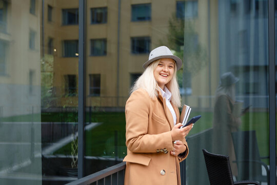 woman in a beige coat and hat holding a notebook and a smartphone