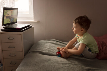 Toddler boy sitting on the bed and watching cartoon on laptop