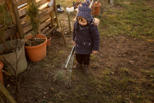 Small boy in winter clothes raking ground with big  and rusty ra