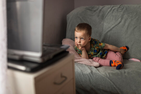 Small blonde boy lying down on sofa and watching cartoons on lap