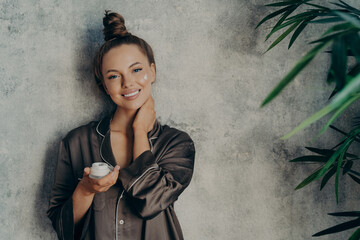 Beautiful caucasian woman in silk pajama with face cream smiling at camera