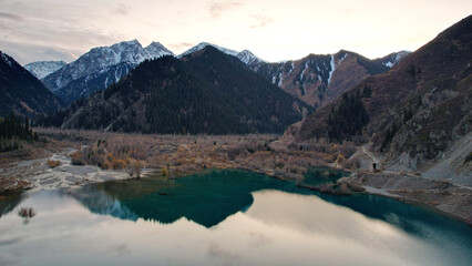 Issyk mountain lake with mirror water at sunset. The color of the water changes before our eyes. There are trees in clear water. Snowy mountains and green hills are visible. Clouds are reflected