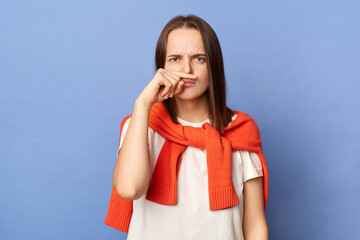 Image of young adult Caucasian woman wearing white T-shirt and orange sweater tied over shoulders standing isolated on blue background, keeps finger under nose, looking at camera.