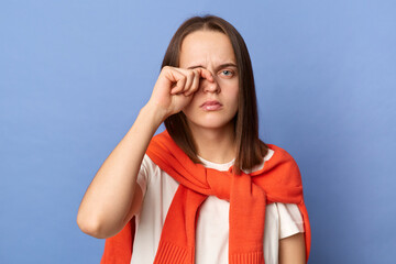 Indoor shot of tired exhausted woman wearing white T-shirt and orange sweater tied over shoulders standing isolated on blue background, looking at camera, rubbing her eye.