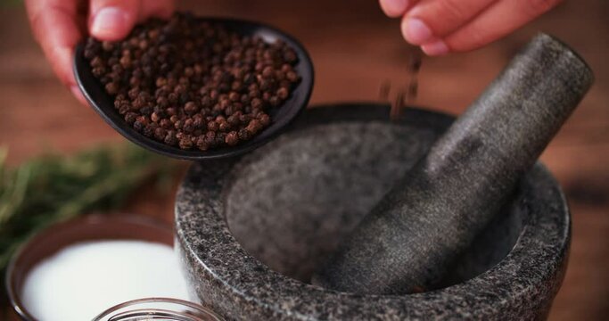 Person's hands putting black peppercorns into a dark stone mortar and pestle for crushing, with herbs and spices laid out on a wooden table in Slow Motion