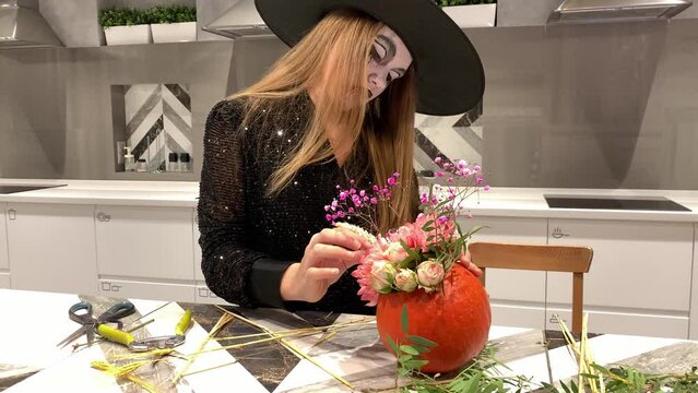 A girl in a witch costume and with a painted face creates a flower composition from a pumpkin. High quality photo