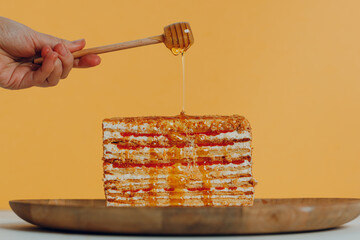 Slice of layered honey cake with stick in hand on plate yellow background.