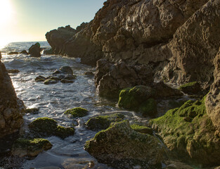 beach or coast of rocky area with waves breaking between the rocks with a sun in the background a calm sea with moss or seaweed on the rocks with room for text