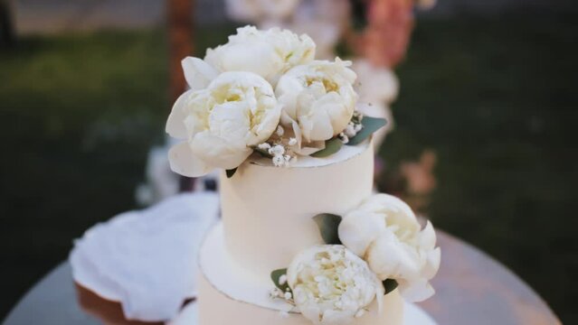 Close-up wedding cake decorated with white peonies flowers slow motion. Beautiful wedding cake white colored, wedding arch on the background.