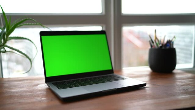 Laptop with green screen in soft focus at workplace. Open computer with chroma key on a wooden table behind a window. Workplace at home. Selective focus.