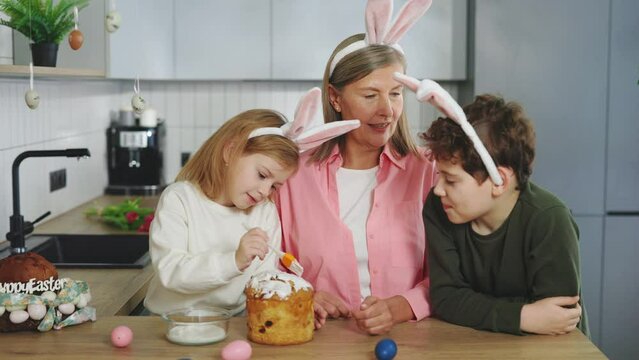 Happy grandmother with grandchildren wearing bunny ears decorating easter cake at home together. Family spending time together. Preparing for the holiday