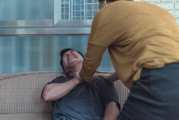 A panicked wife grabs the hand of her husband who is experiencing a heart attack while sitting on the sofa in the living room.
