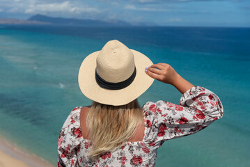 Mujer rubia con el pelo largo en la espalda sosteniendo un sombrero de paja frente al mar turquesa...