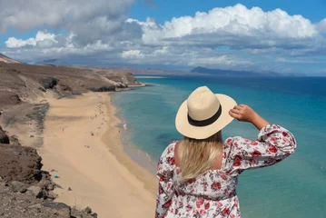 Papier Peint photo autocollant Plage de Sotavento, Fuerteventura, Îles Canaries Mujer rubia con el pelo largo en la espalda sosteniendo un sombrero de paja frente al mar turquesa y la playa de arena blanca rodeada de un paisaje volcánico en Fuerteventura, Islas Canarias.