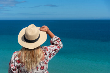 Mujer rubia con el pelo largo en la espalda sosteniendo un sombrero de paja frente al mar turquesa...