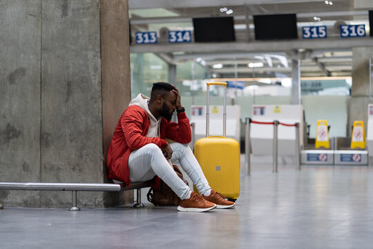Sad African American man upset at airport his flight is delayed. Depressed traveler male waiting for a plane sitting in empty terminal with baggage. Exhausted guy on a long night connection at airport