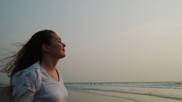 Beautiful young woman with long hair flowing on the wind. Happy female traveller enjoys freedom on the seashore at sunset.