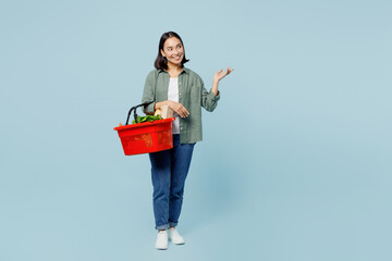Full body young woman in casual clothes hold red basket with food products point index finger aside...