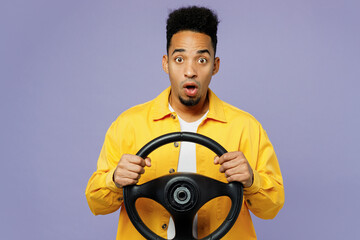 Young puzzled surprised man of African American ethnicity wear yellow shirt t-shirt hold steering wheel driving car isolated on plain pastel light purple background studio. People lifestyle concept.