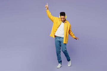 Full body young man of African American ethnicity wearing yellow shirt t-shirt headphones listen music dance raise up hands isolated on plain pastel light purple background People lifestyle concept.