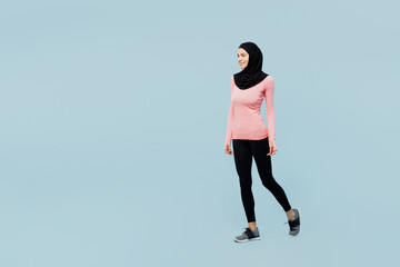 Full body smiling young asian muslim fitness trainer sporty woman wears pink abaya hijab spend time in home gym walking going isolated on plain blue background studio. Workout sport fit abs concept.
