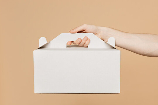 Close up cropped male holding in hand hold cake dessert in unmarked clear empty blank white cardboard box isolated on pastel plain light beige color wall background studio. Service concept concept