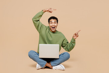 Full body young man of African American ethnicity wear green sweatshirt sit hold use work on laptop pc computer point aside isolated on plain pastel light beige background People lifestyle concept.