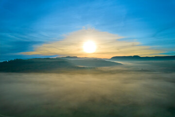Aerial view of foggy landscape at sunrise. Mountains in front of valley with yellow sun in autumn. Germany, Nurtingen, Tiefenbachtal.