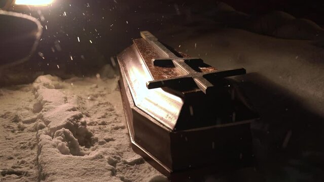 A closed coffin lies on the ground in the snow at night. Strong blizzard. Burning car headlights