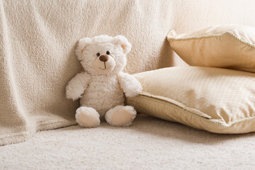 Smiling white teddy bear sitting on carpet at sofa and pillows at nursery room. Closeup. Front view.