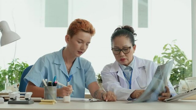 Young female clinician making notes in medical document while listening to experienced colleague explaining patient lung x-ray result during their meeting in hospital