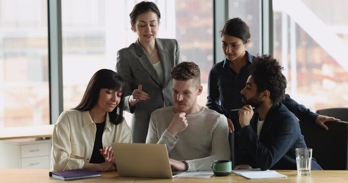 Engaged business mentor woman training interns at laptop, explaining professional app, software work, standing at computer, speaking to diverse group of employees